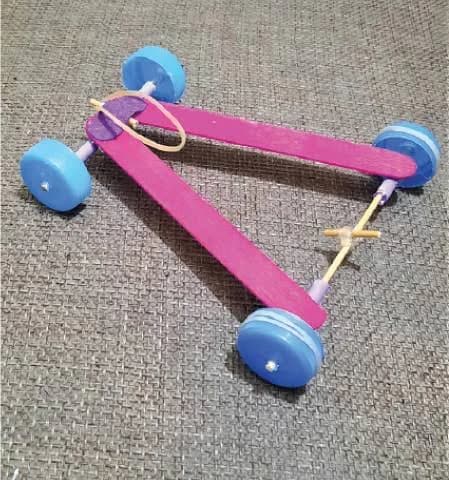 A small pink craft-stick car with four blue wheels and a visible rubber-band drive.